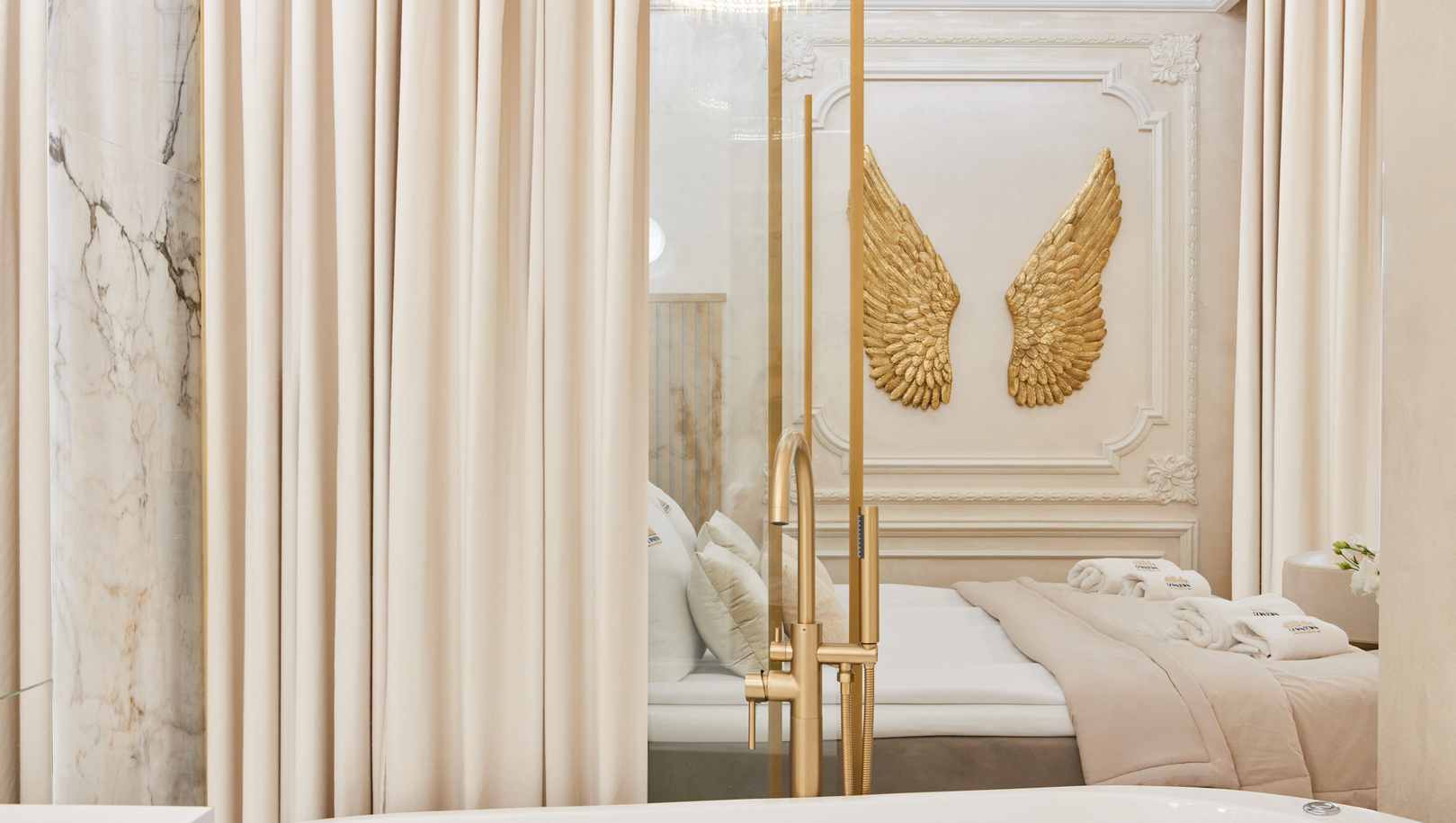 Biała wolnostojąca wanna i złoty kran, marmur i zasłony; widok na przytulne łóżko z rulonami ręczników i złotymi skrzydłami.