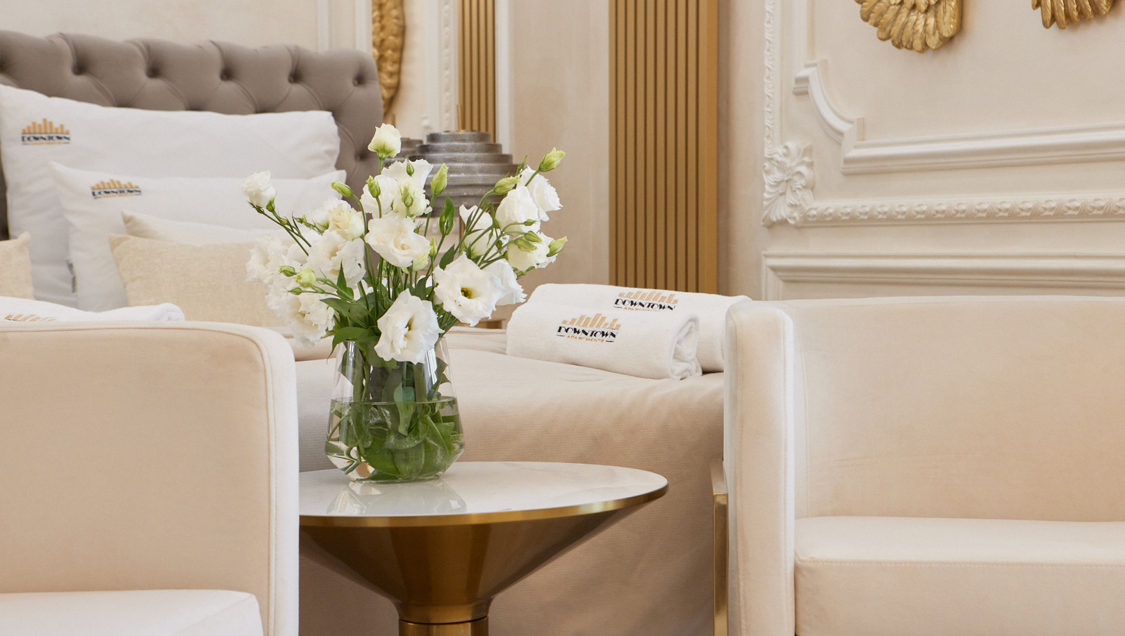 Luksusowa strefa w apartamencie: kremowe fotele, złoty stolik z wazonem białych kwiatów, haftowane ręczniki na łóżku