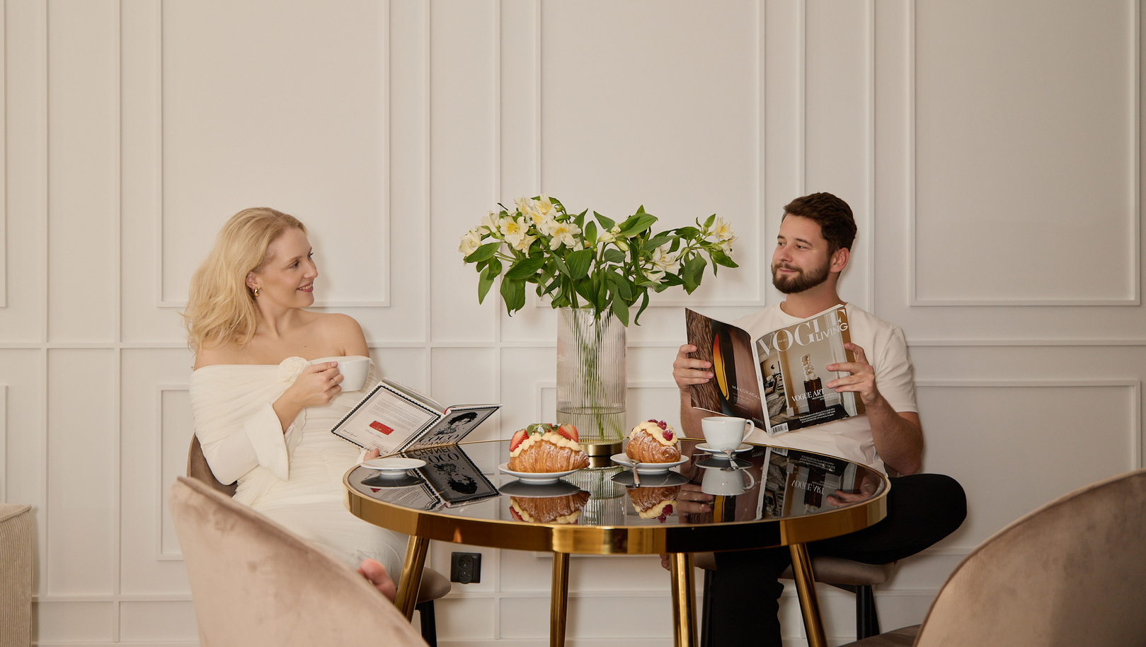 Para przy hotelowym śniadaniu: kawa i croissanty z owocami na złotym stole, świeże kwiaty i wygodne fotele.