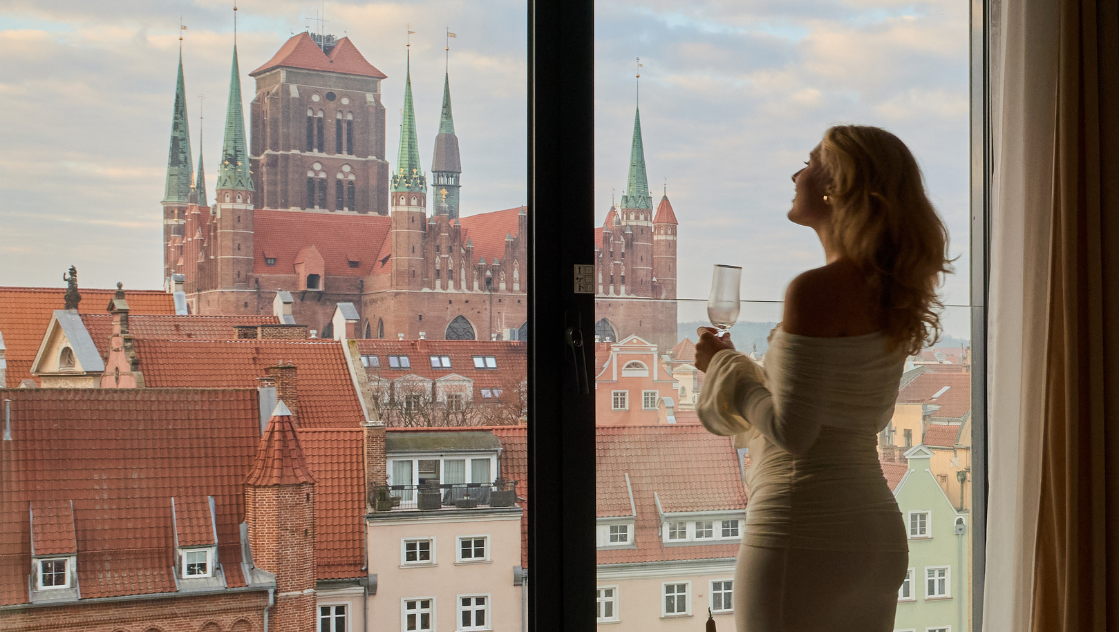Gość w hotelowym szlafroku trzyma kieliszek wina przy panoramicznym oknie, widok na dachy i wieże katedry