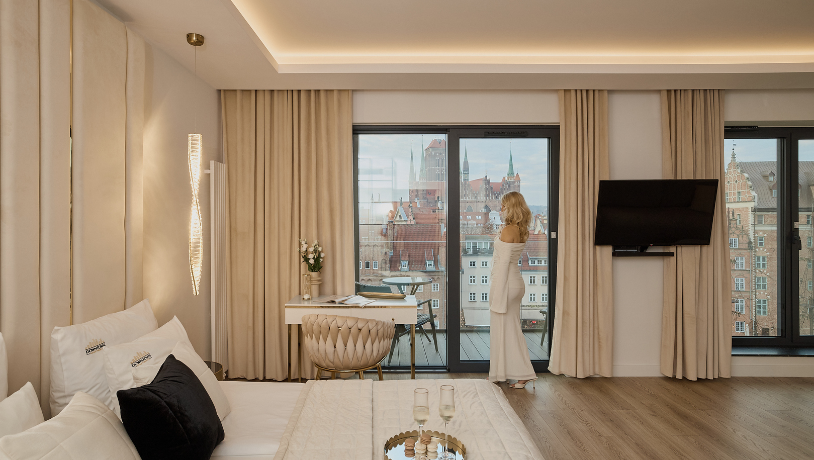 Apartament z widokiem na miasto, duże łóżko, beżowe wnętrze, biurko i balkon z dachami; gość w eleganckiej sukni.