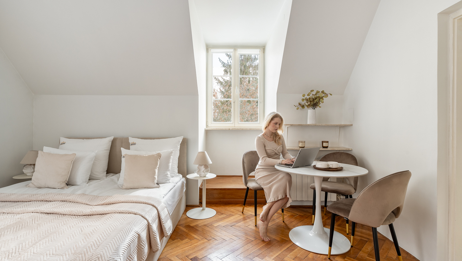 Jasny pokój na poddaszu z dużym łóżkiem, drewnianą podłogą jodełką, okrągłym stolikiem; gość pracuje na laptopie przy oknie.
