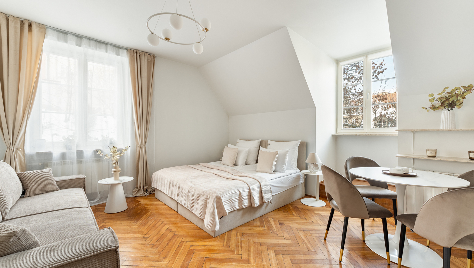 Jasne studio z łóżkiem małżeńskim, sofą, stołem jadalnym, podłogą w jodełkę i dużymi oknami w neutralnej kolorystyce.