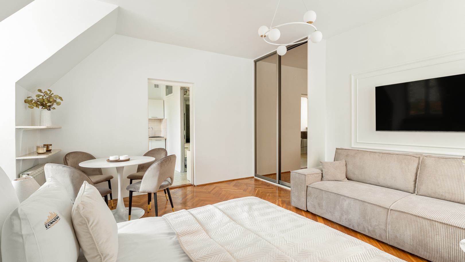 Jasny, nowoczesny studio z dużym łóżkiem, wygodną sofą, telewizorem, stołem dla czterech, lustrem i parkietem.