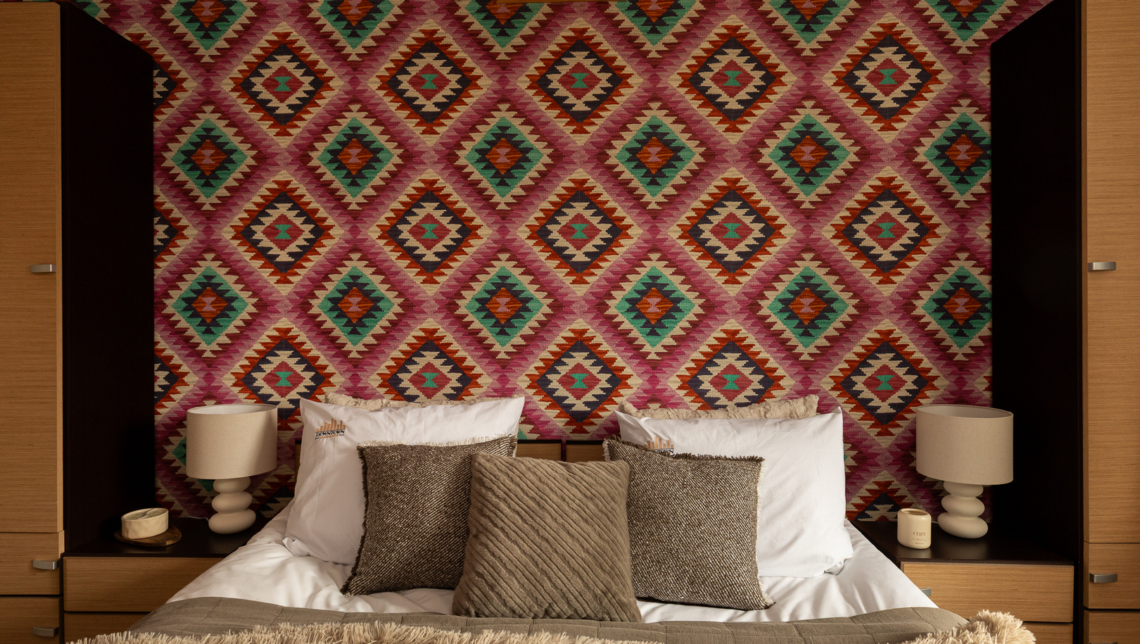 Podwójne łóżko z kilkoma poduszkami i miękką narzutą przed kolorową, geometryczną ścianą; drewniane stoliki i lampki