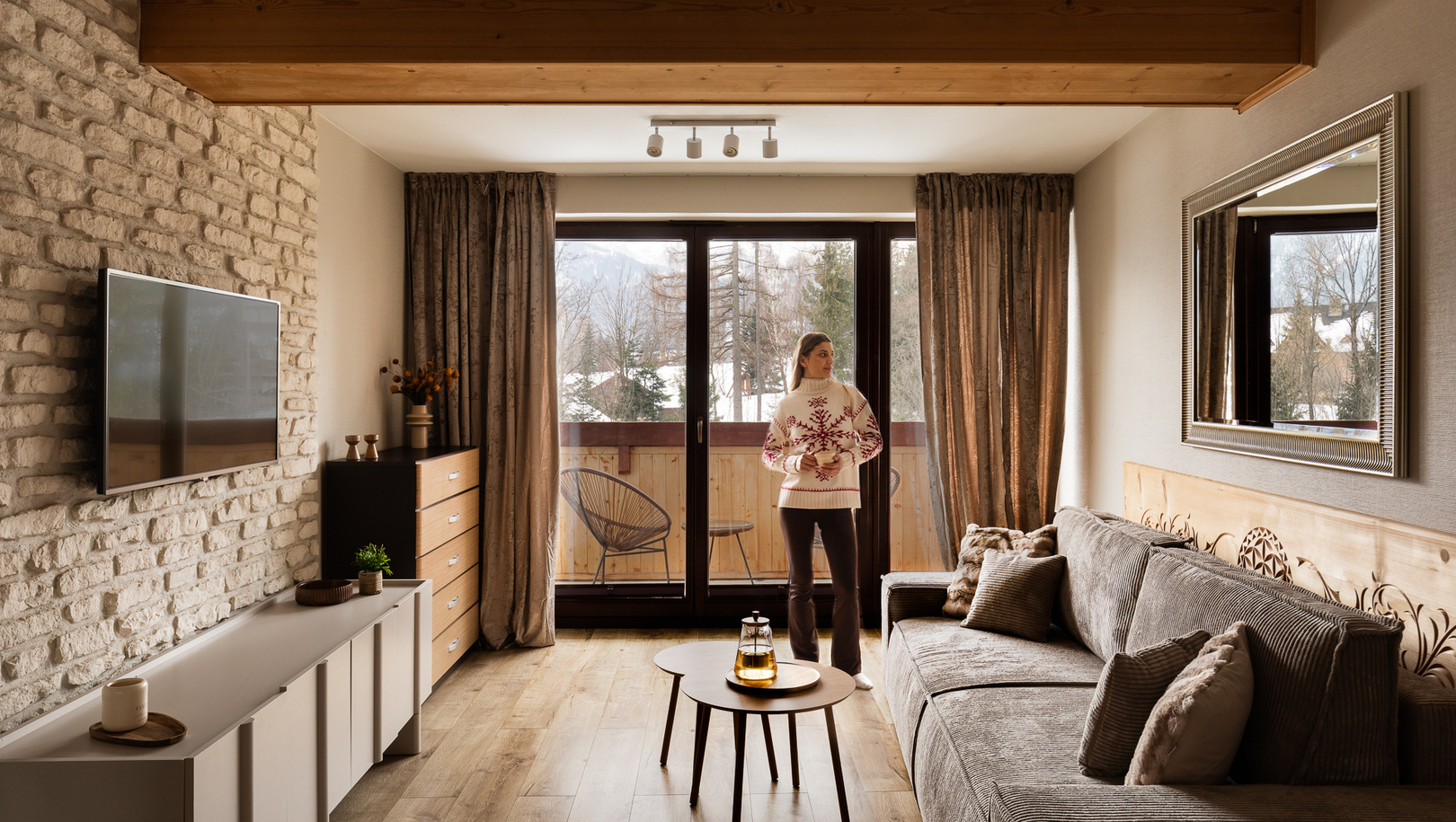 Przytulny salon w górskim apartamencie z sofą, drewnianymi belkami i balkonem; gość z gorącym napojem i widokiem na śnieg.