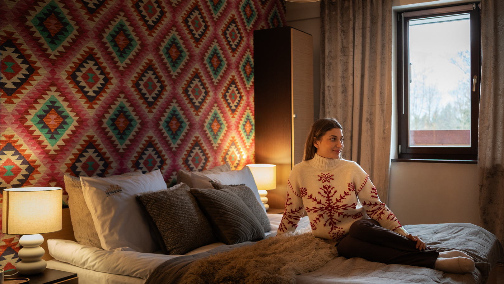 Przytulny hotelowy pokój z wzorzystą ścianą, miękką pościelą, lampkami nocnymi i kobietą w swetrze przy oknie.