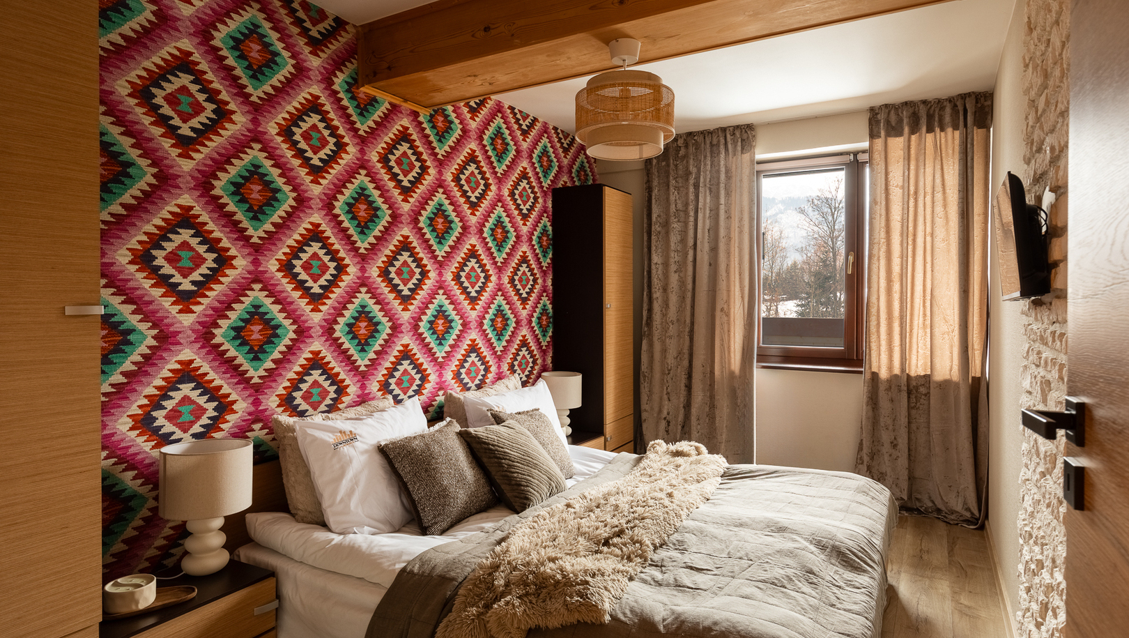Przytulny pokój dwuosobowy z kolorową geometryczną ścianą, drewnianą belką, miękką pościelą i widokiem na góry.