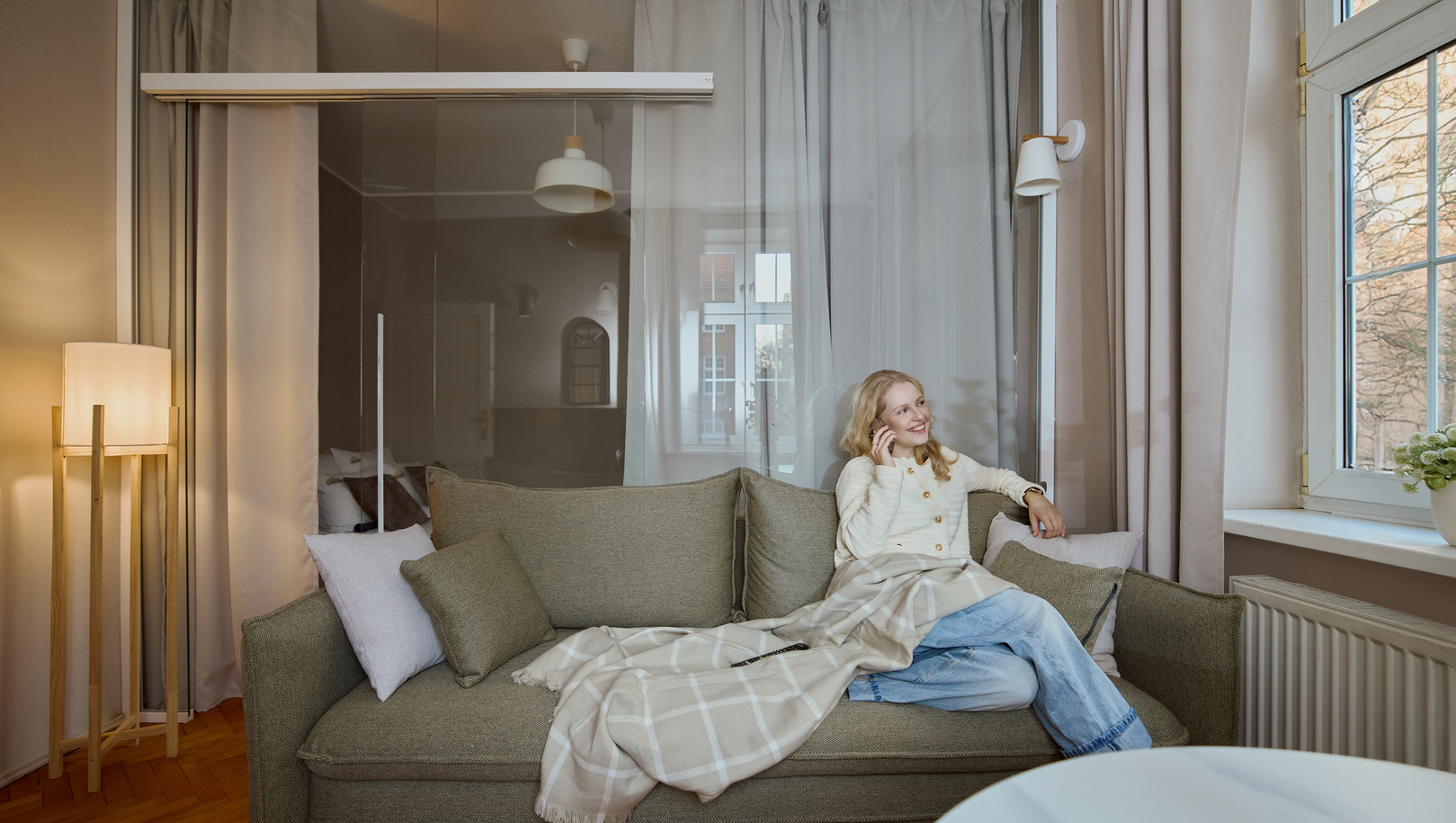 Uśmiechnięta kobieta na sofie z kocem i poduszkami w przytulnym apartamencie, rozmawia przez telefon przy dużym oknie.