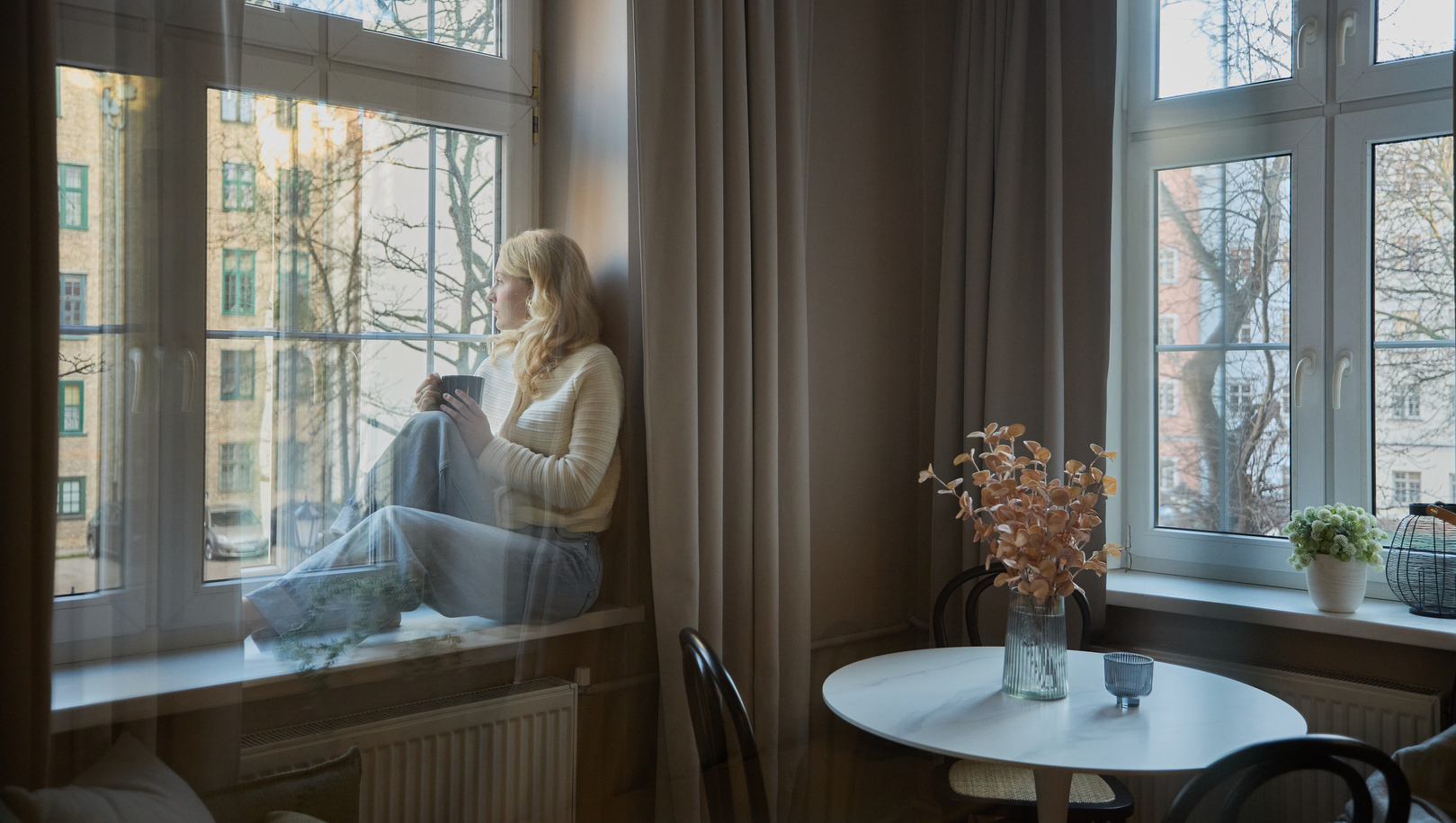 Przytulne miejskie studio z kobietą na siedzisku przy oknie trzymającą kubek, okrągły stolik z wazonem, duże okna, zasłony i widok na kamienice