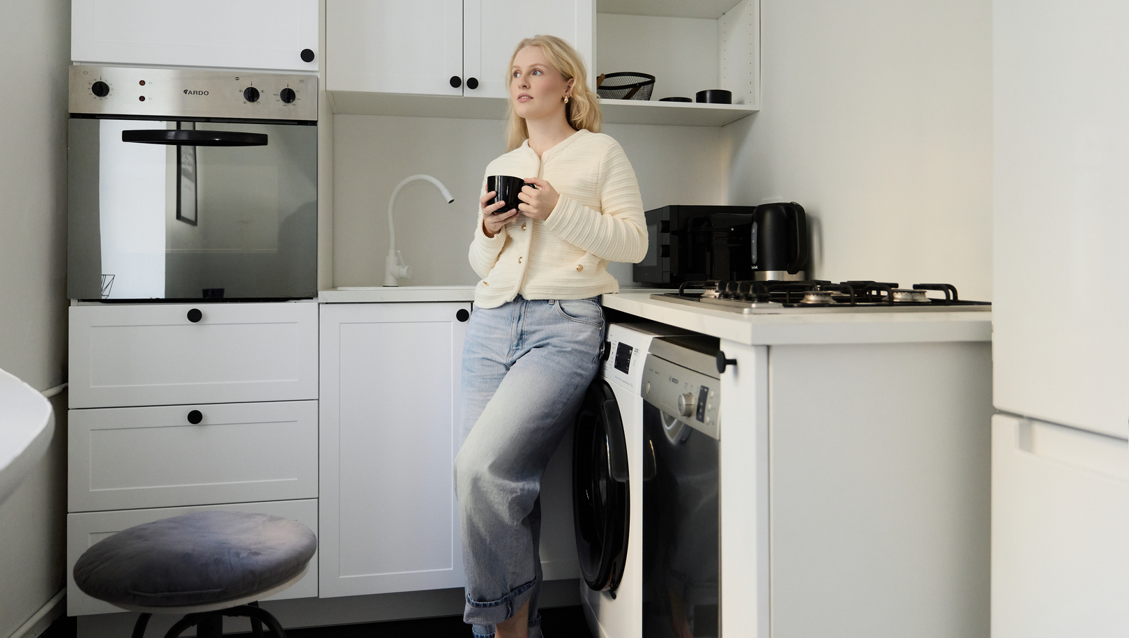 Kobieta oparta o biały aneks kuchenny w nowoczesnym apartamencie, trzyma czarny kubek; płyta gazowa, piekarnik i pralka.