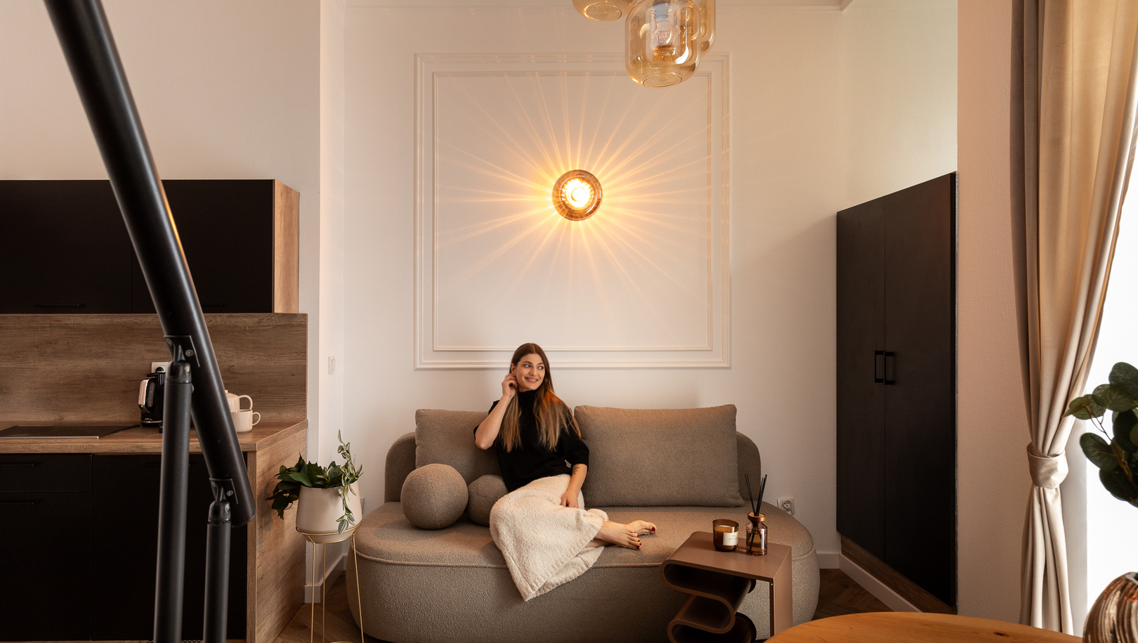 Przytulna część dzienna w studio z zaokrągloną sofą, dekoracyjną lampą ścienną, aneksem kuchennym i kobietą odpoczywającą pod kocem