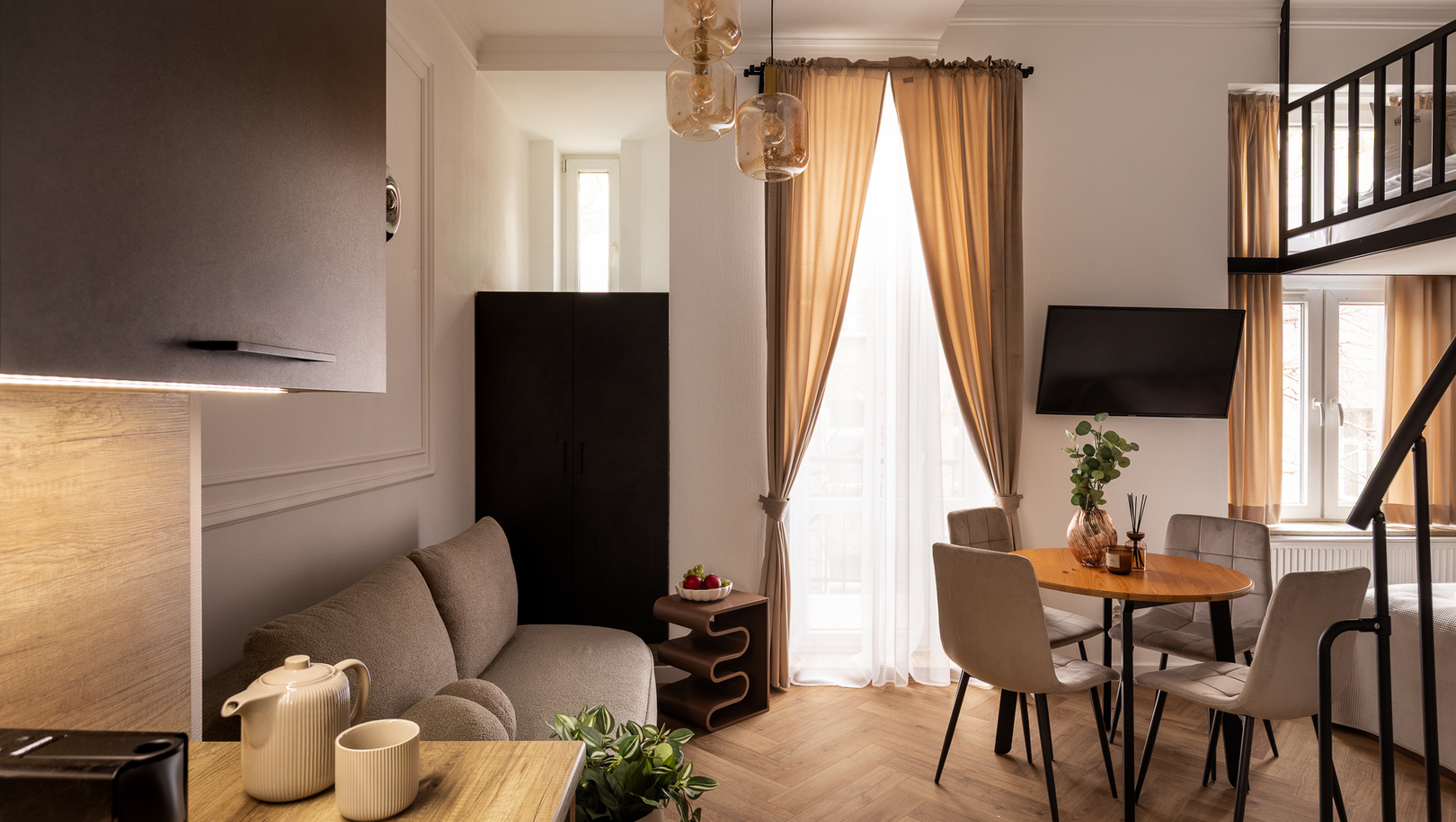 Przytulne, nowoczesne studio z antresolą, stołem jadalnym, sofą, telewizorem, czajnikiem w aneksie i drewnianą podłogą