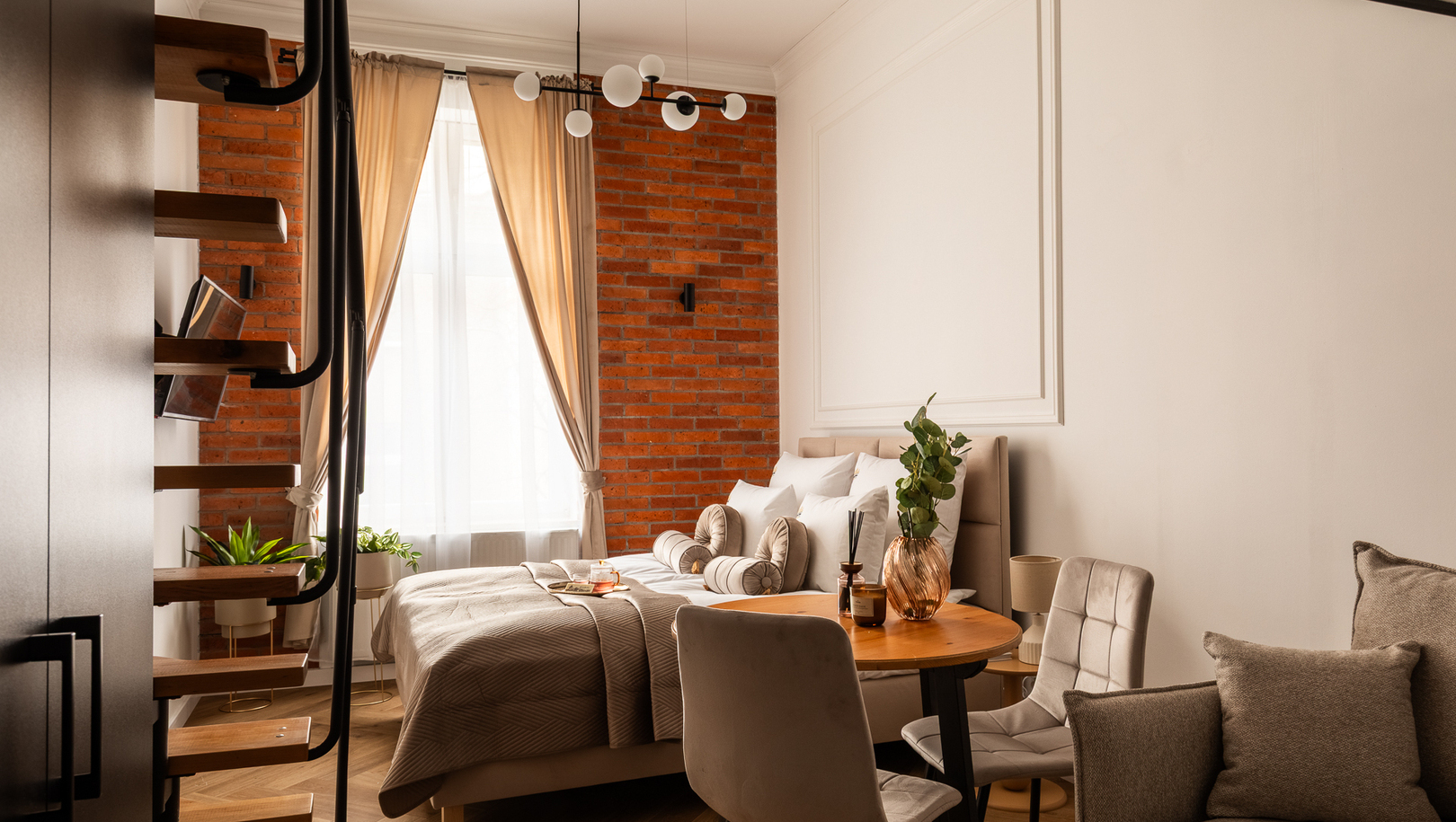 Przytulne studio z dużym łóżkiem, ceglanym murem, beżową pościelą, okrągłym drewnianym stołem, lampą wiszącą i roślinami.