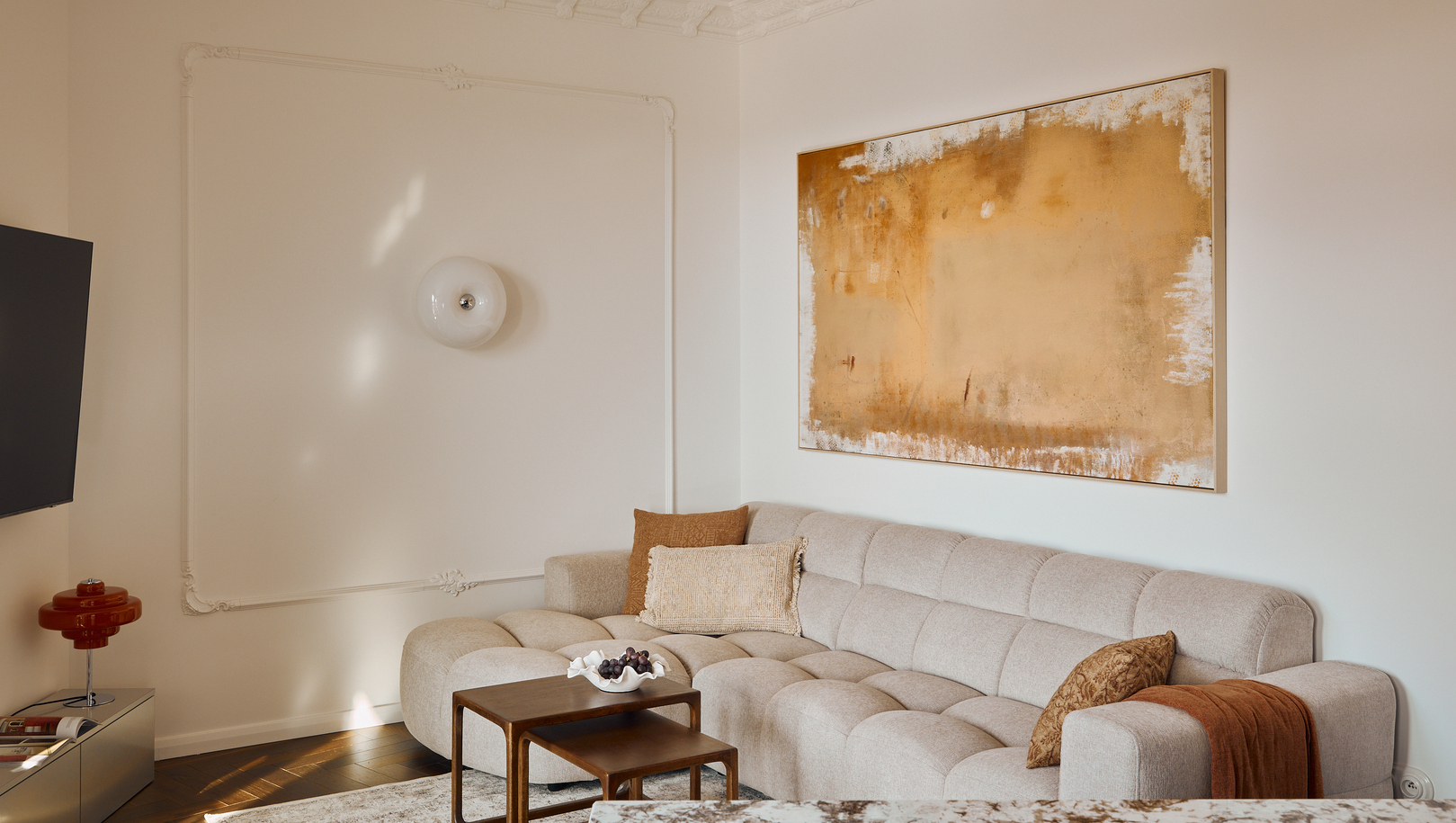 Jasny salon w apartamencie hotelowym z miękką pikowaną sofą narożną, dużym złotym obrazem, marmurowym blatem i drewnianymi stolikami.