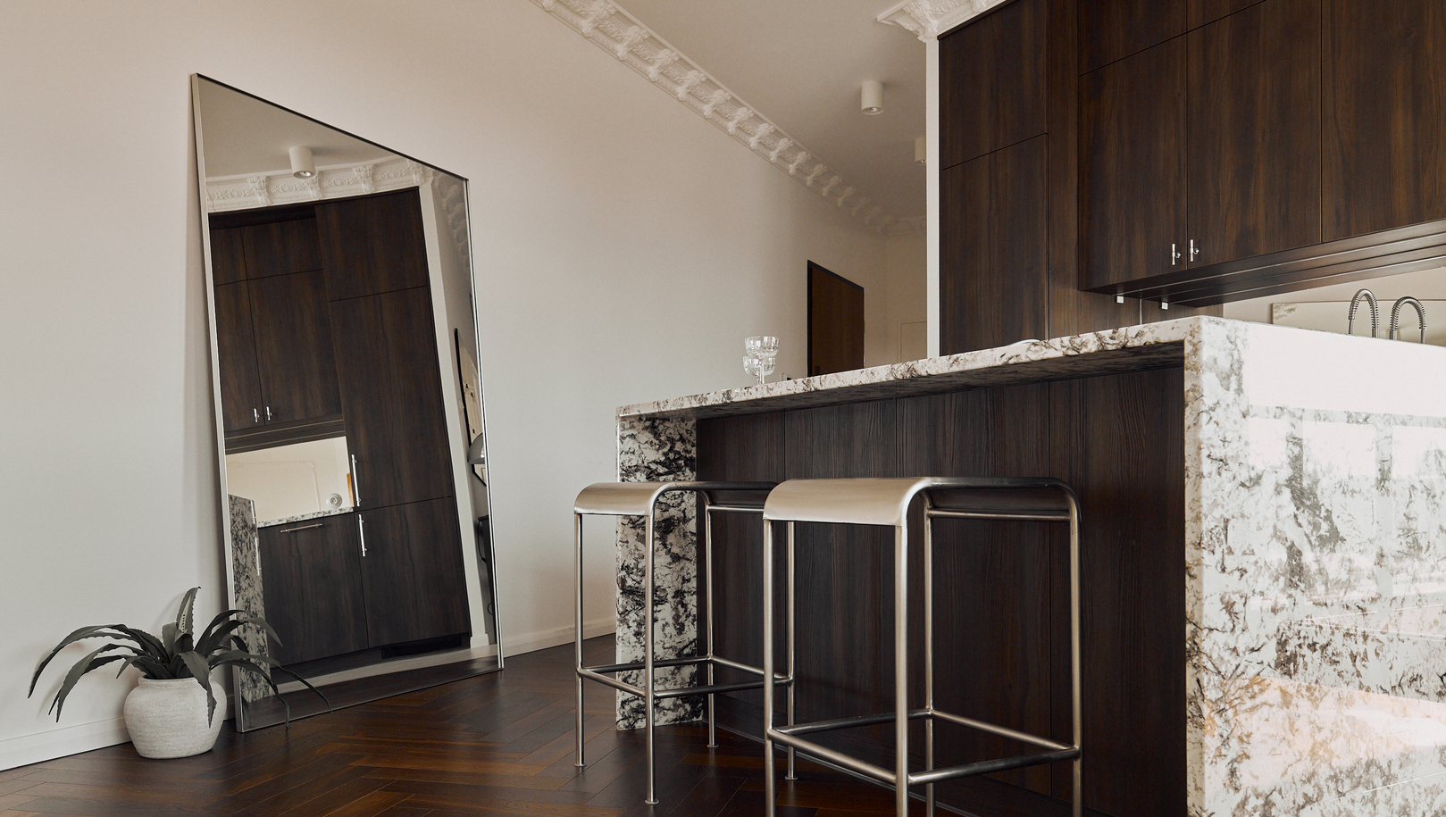Nowoczesny aneks kuchenny w apartamencie z marmurową wyspą, dwoma metalowymi stołkami, ciemnymi szafkami i dużym lustrem