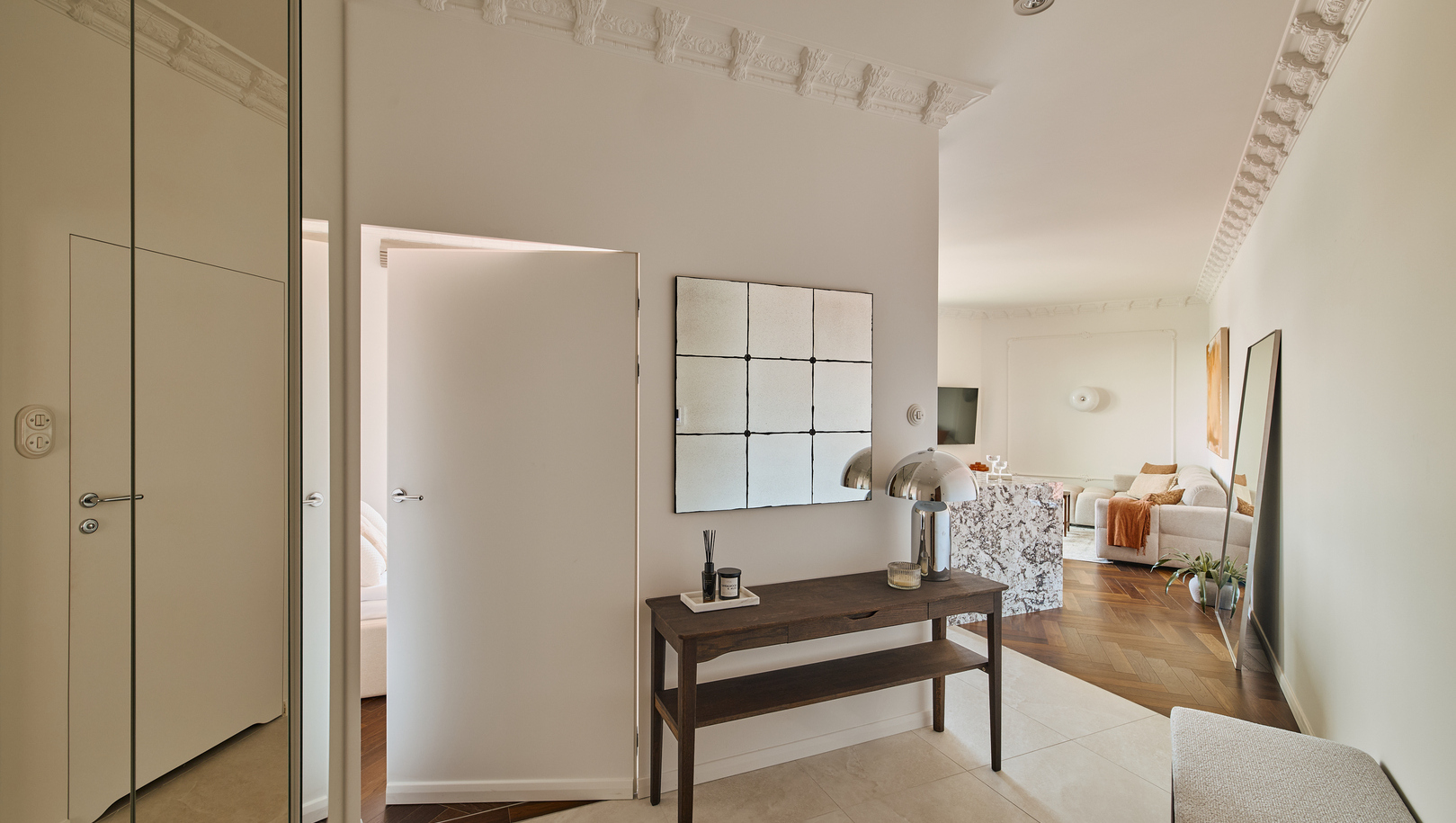 Przedsionek z drewnianą konsolą, lustrzaną mozaiką, chromowaną lampą, ozdobnym gzymsem i widokiem na jasny salon.