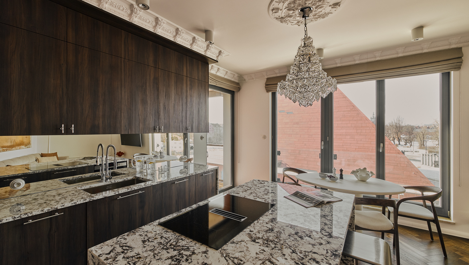 Kuchnia w luksusowym apartamencie z marmurową wyspą i płytą grzewczą, ciemnymi drewnianymi szafkami, kryształowym żyrandolem i widokiem na miasto.