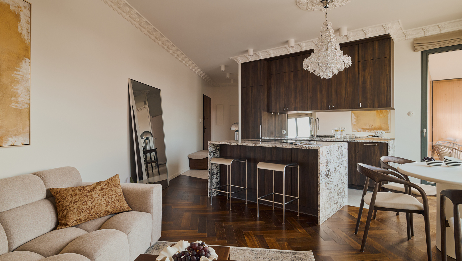 Apartament z otwartą kuchnią, marmurową wyspą, ciemnymi drewnianymi szafkami, kryształowym żyrandolem, wygodną sofą i parkietem jodełką