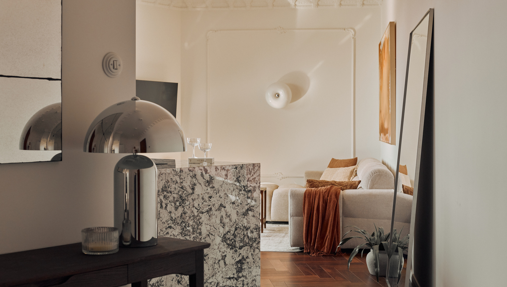 Elegancki salon apartamentu z marmurową ladą, kremową sofą z rdzawym pledem, dużym lustrem opartym o ścianę, chromowaną lampą i parkietem