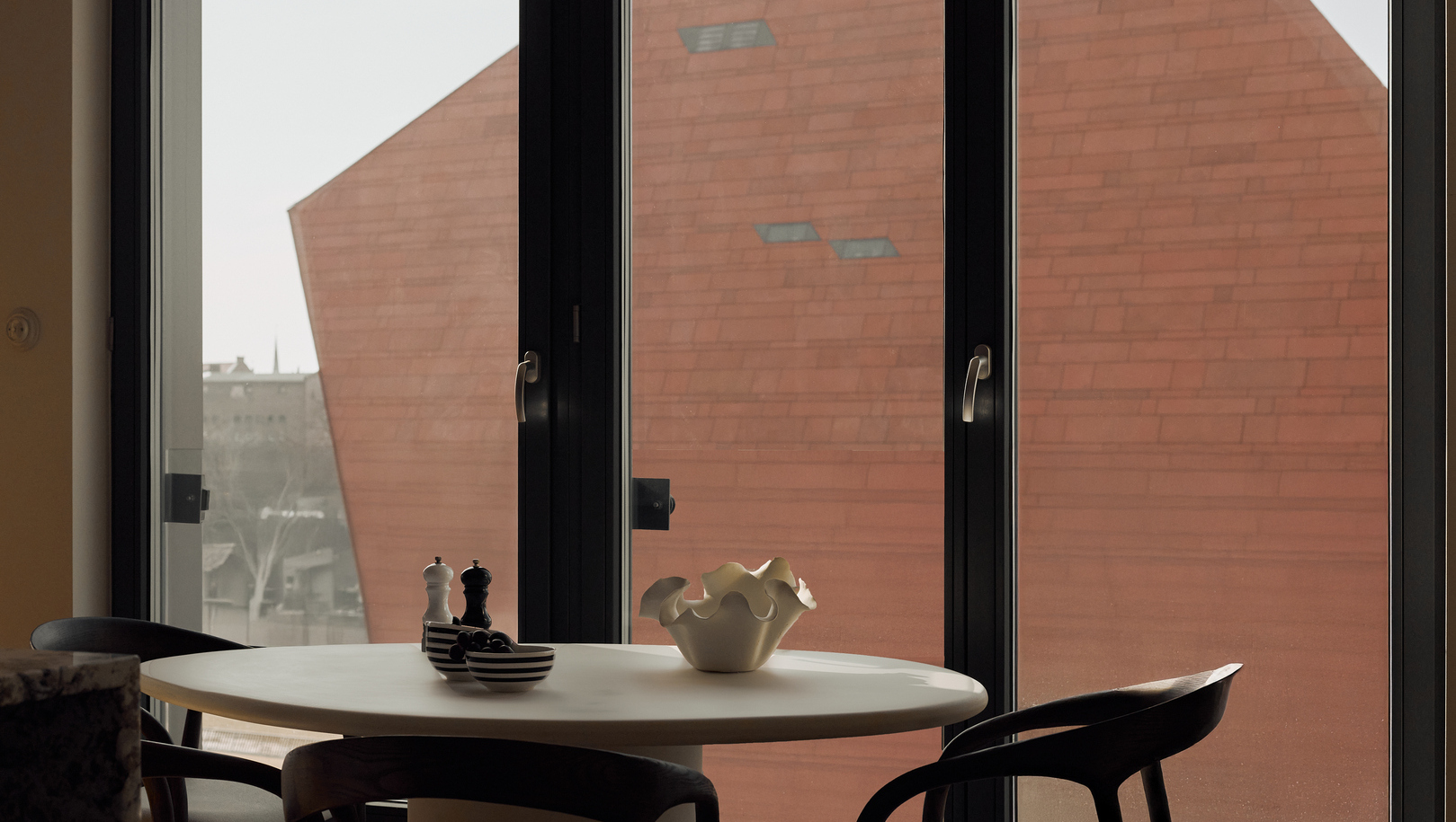 Kameralny kącik jadalny w hotelu z okrągłym białym stołem, rzeźbiarską ozdobą, paskowanymi miseczkami i drewnianymi krzesłami przy wysokich oknach i czerwonej fasadzie