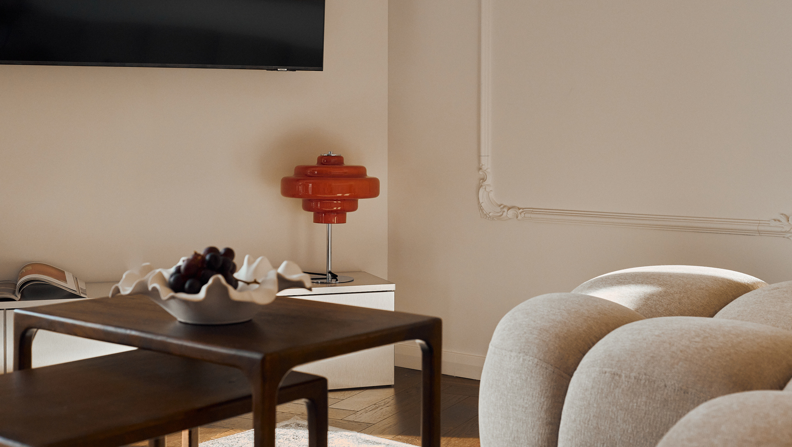 Przytulna, zaokrąglona sofa, drewniane stoliki nido z dekoracyjną misą winogron, czerwona lampa stołowa, ozdobne listwy i telewizor na ścianie