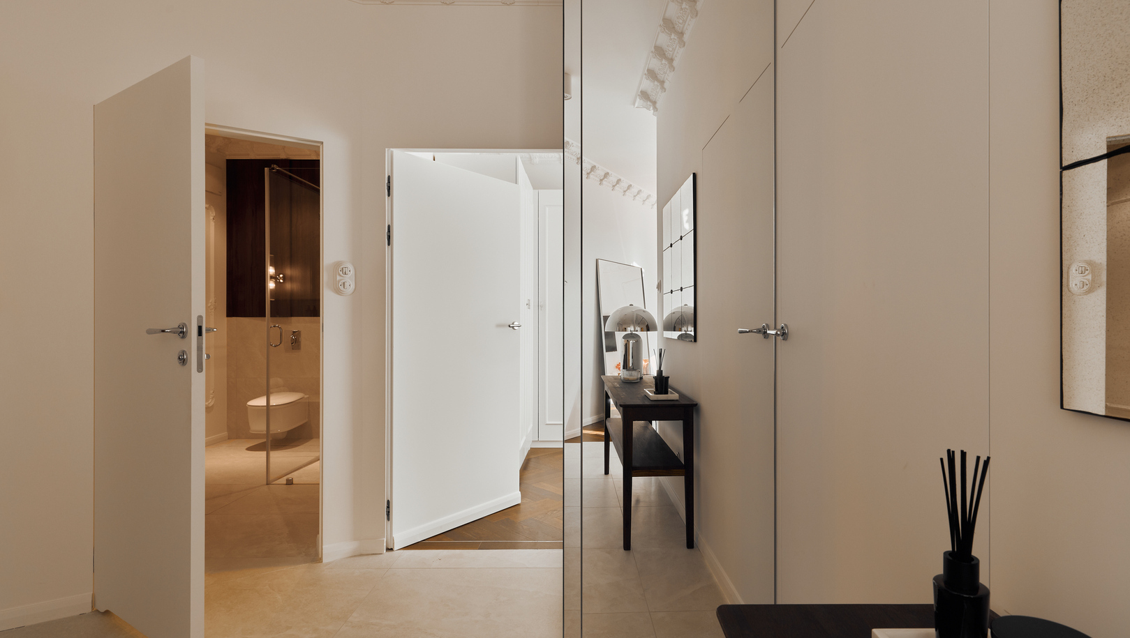 Elegancki hol z lustrzaną szafą, drewnianą konsolą z lampą i dyfuzorem, otwarte drzwi do nowoczesnej marmurowej łazienki i dekoracyjny sufit.