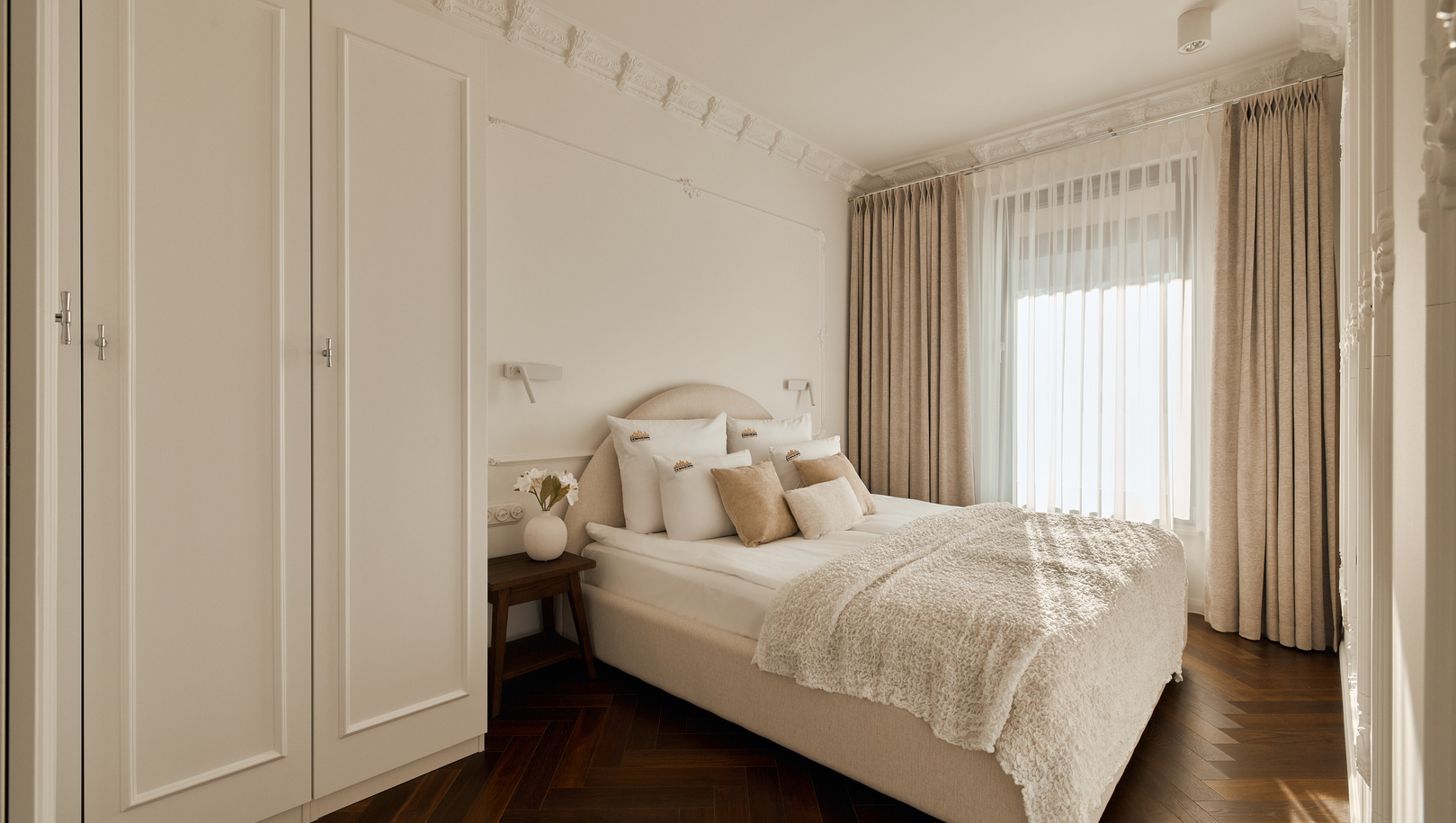 Elegancki pokój dwuosobowy z beżowym łóżkiem, puszystą narzutą, ozdobnymi listwami sufitowymi i parkietem