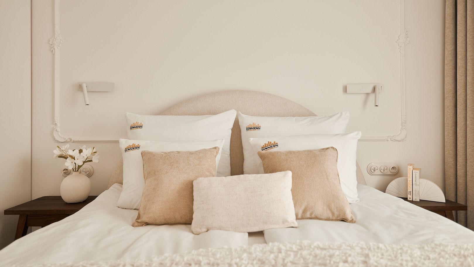 Łóżko dwuosobowe w neutralnych tonach z zaokrąglonym zagłówkiem, białą pościelą, beżowymi poduszkami, lampkami, wazonem i dekoracyjnym stiukiem.