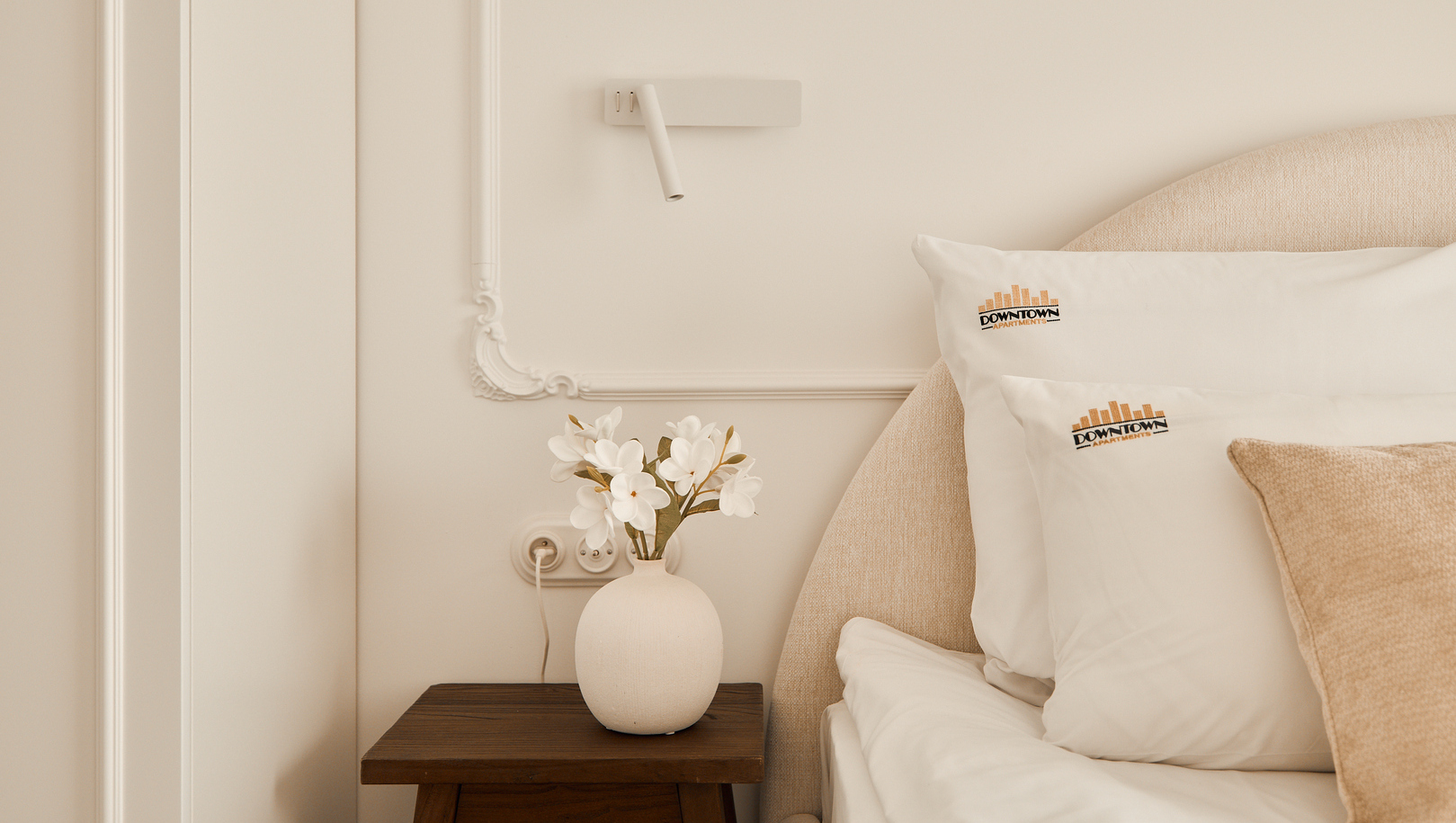 Butikowa sypialnia z tapicerowanym zagłówkiem, poduszkami z logo 'Downtown Apartments', stolik i wazon z białymi kwiatami.