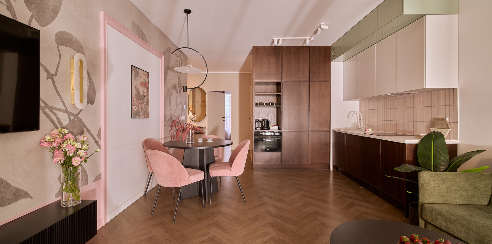 Aneks apartamentu z jadalnią: okrągły stół, różowe aksamitne krzesła, jodełkowa podłoga, sofa, telewizor i nowoczesne AGD.