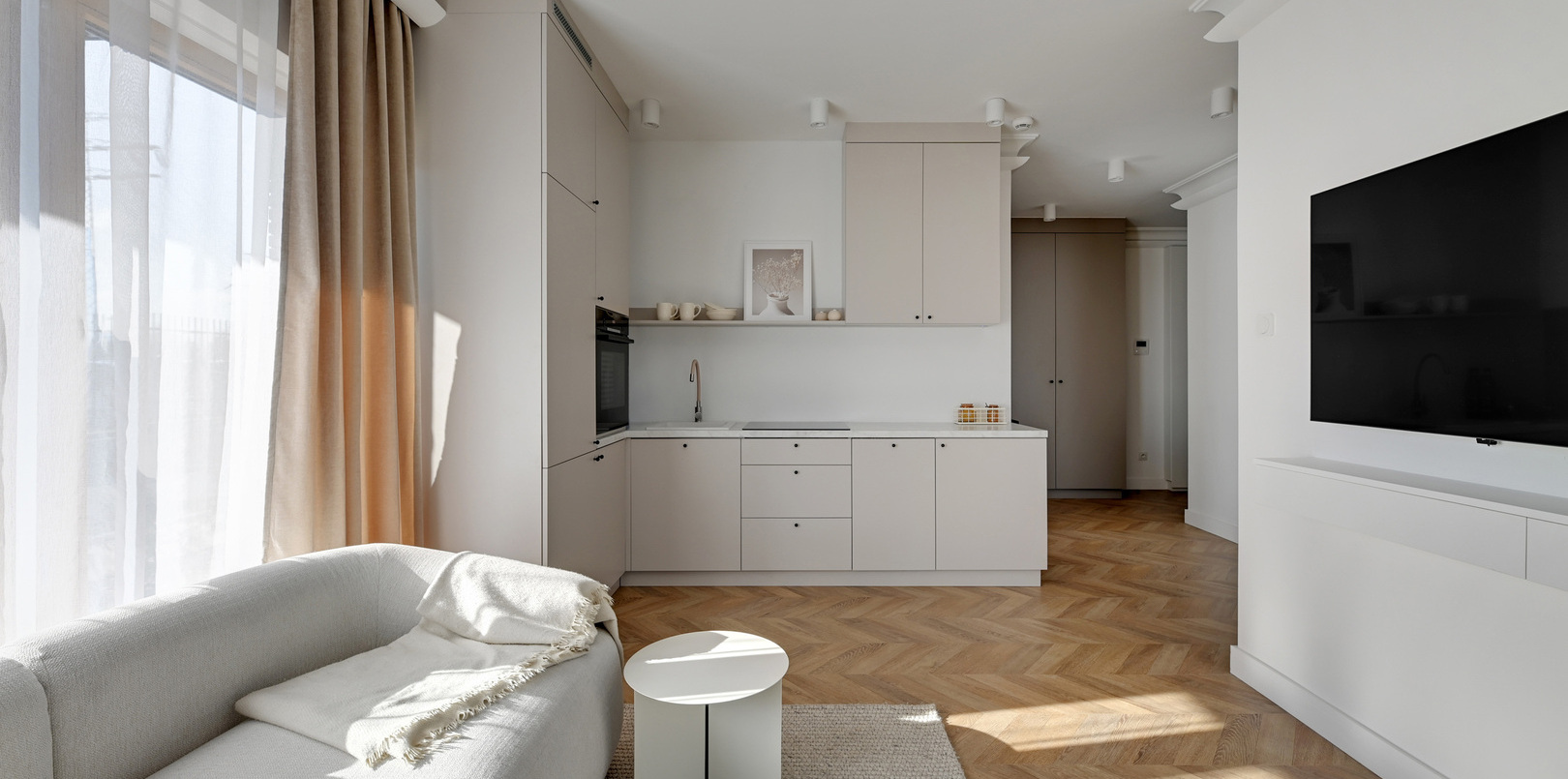 Jasne studio z aneksem kuchennym, sofą, małym okrągłym stolikiem, TV, zasłonami do podłogi i parkietem w jodełkę.