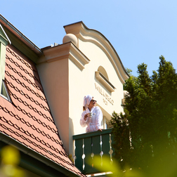 Gość w szlafroku na słonecznym balkonie willi, pijący kawę przy ozdobnej elewacji i czerwonym dachu, otoczony drzewami