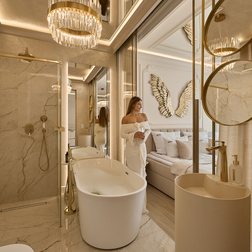 Łazienka z marmurem, wolnostojącą wanną, złotymi armaturami i żyrandolem, otwierająca się na sypialnię ze złotymi skrzydłami; kobieta w białym szlafroku z drinkiem.