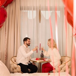 Para wznosząca toast szampanem w pokoju hotelowym udekorowanym czerwonymi balonami w kształcie serc, płatkami róż i świecami.