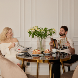 Para pijąca poranną kawę i jedząca wypieki przy złoconym okrągłym stoliku w eleganckim apartamencie, wazon z liliami