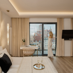 Luksusowy apartament z widokiem na miasto, duże łóżko, beżowe wnętrze, biurko i balkon; kobieta w białej sukni patrzy na zabytkowe dachy i wieże; taca z dwoma kieliszkami szampana