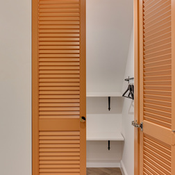 Pomarańczowe drzwi żaluzjowe otwierające się na białą garderobę w pokoju z półkami, wieszakami i podłogą w jodełkę