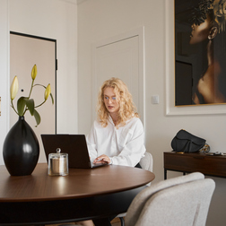 Jasna nowoczesna część jadalna w apartamencie z okrągłym drewnianym stołem, czarnym wazonem z liliami i gościem przy laptopie.