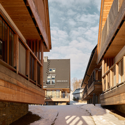 Drewniane chaty z balkonami przy zaśnieżonej ścieżce prowadzącej do budynku ze stromym dachem, przytulna alpejska atmosfera.