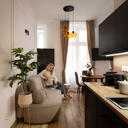 Przytulne studio z aneksem kuchennym, drewnianym blatem i bursztynowymi lampami; gość na sofie pije kawę i czyta.