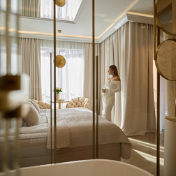 Luksusowy apartament z dużym łóżkiem, pikowanymi fotelami i stolikiem z kwiatami, wolnostojąca wanna i złote baterie, kobieta w białej sukni patrzy w światło.