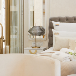 Łóżko z pikowanym zagłówkiem, miękka biała pościel, lampa przy stoliku, złota bateria wolnostojąca, świeże kwiaty i zwinięte ręczniki