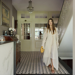 Uśmiechnięta kobieta w zwiewnej sukni przechodzi przez eleganckie wejście hotelu butikowego z wzorzystymi kaflami, żyrandolem i schodami.