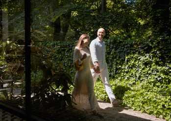 Para idąca za rękę po słonecznej ścieżce hotelowego ogrodu przy szklanej fasadzie i ścianie porośniętej bluszczem