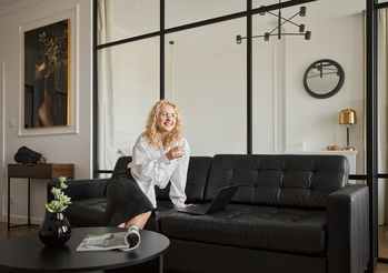 Gość relaksuje się na czarnej skórzanej sofie w apartamencie, otwarty laptop, trzyma szklankę, szklana przegroda i nowoczesne wnętrze.