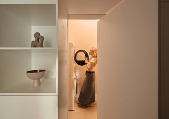 Gość wkłada pranie do pionowej pralko-suszarki w ciepłej, minimalistycznej pralni hotelowej z dekoracyjną półką.