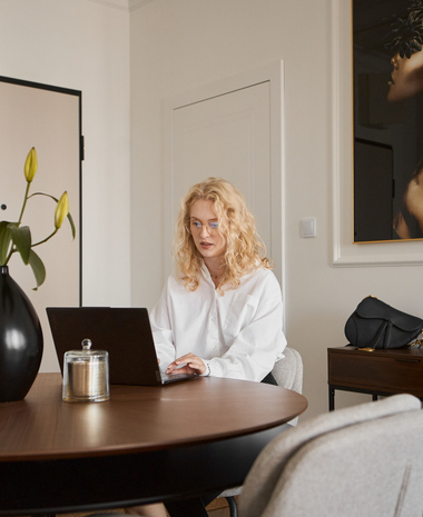 Nowoczesny kącik w apartamencie z okrągłym drewnianym stołem, wazonem z liliami i świecą, kobieta pracuje przy laptopie, elegancka grafika