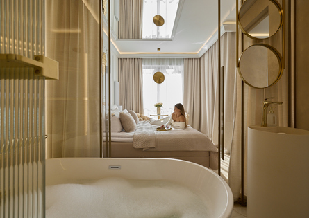 Luksusowy apartament z wolnostojącą wanną pełną piany, neutralne odcienie i złote akcenty; kobieta relaksuje się na łóżku z tacą śniadaniową.