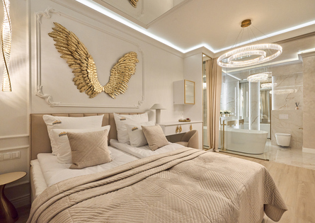 Elegancki apartament z beżową pikowaną pościelą, złotą dekoracją w kształcie skrzydeł, przeszkloną łazienką z wolnostojącą wanną i kryształowym żyrandolem